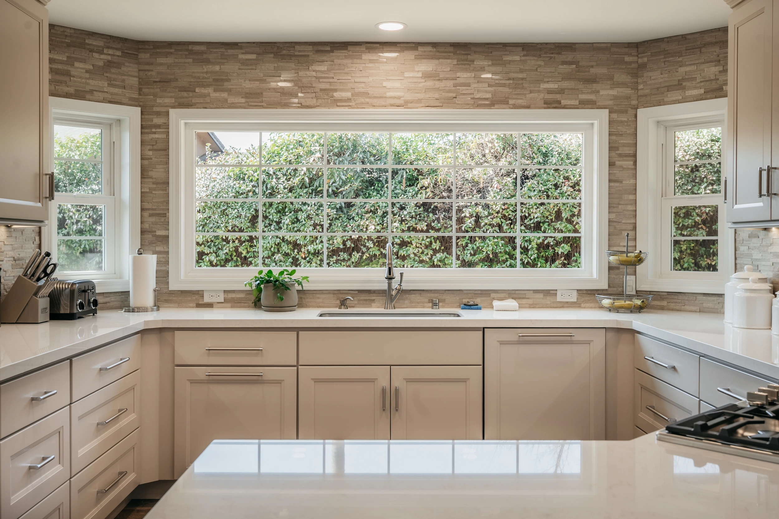 A modern kitchen with beige cabinets, white countertops, a large window above the sink showing greenery outside, and small appliances and containers neatly arranged on the counters.