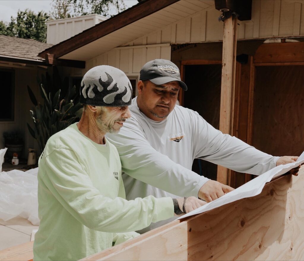 Two men stand at a construction site, examining blueprints on a wooden surface—planning their next Bathroom Remodeling Fresno project. One wears a flame-pattern beanie and green shirt; the other sports a gray cap and light gray long-sleeve shirt.
