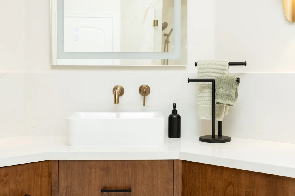 A modern bathroom countertop, ideal for Bathroom Remodeling Fresno projects, features a white rectangular sink, wall-mounted brass faucet, black soap dispenser, and a black stand with two green towels atop light wood cabinetry.