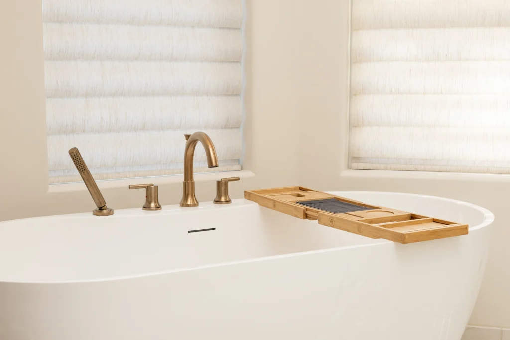 A modern white bathtub with gold fixtures, a handheld shower, and a wooden bath tray sits between two windows with white blinds. This clean, minimal space showcases expert Bathroom Remodeling Fresno style.