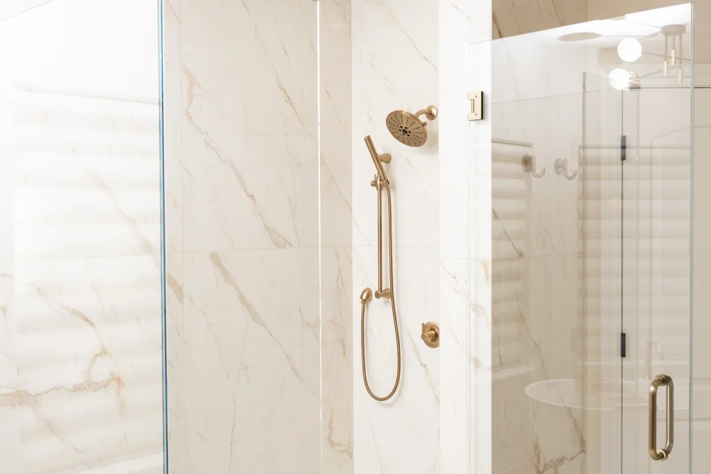 A modern shower perfect for bathroom remodeling in Fresno, featuring beige marble-patterned walls, a glass door, and a gold-colored showerhead with matching handheld attachment and controls.