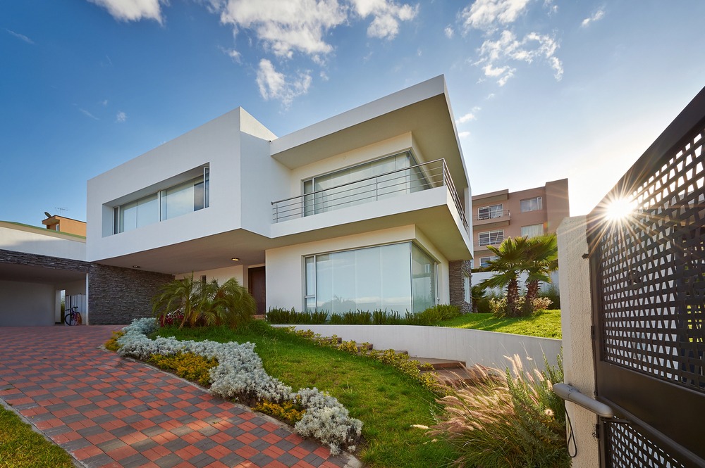Modern two-story white house with large windows, a balcony, and a landscaped front yard. The driveway is paved with red and gray bricks. The sky is blue with some clouds and the sun is shining.