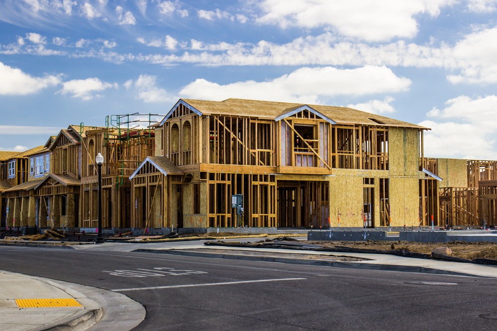 A two-story house under construction, showing exposed wooden framing and roof structure—an example of what to expect when considering a second story addition cost—located at the corner of a paved street under a partly cloudy sky.