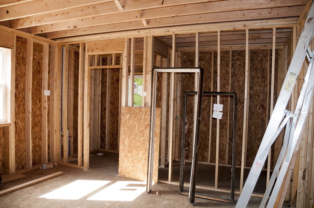 Wooden framing of a house under construction, showing exposed beams, studs, and plywood. A ladder leans against the wall, and window frames are propped up inside the unfinished room. Sunlight comes through a window.