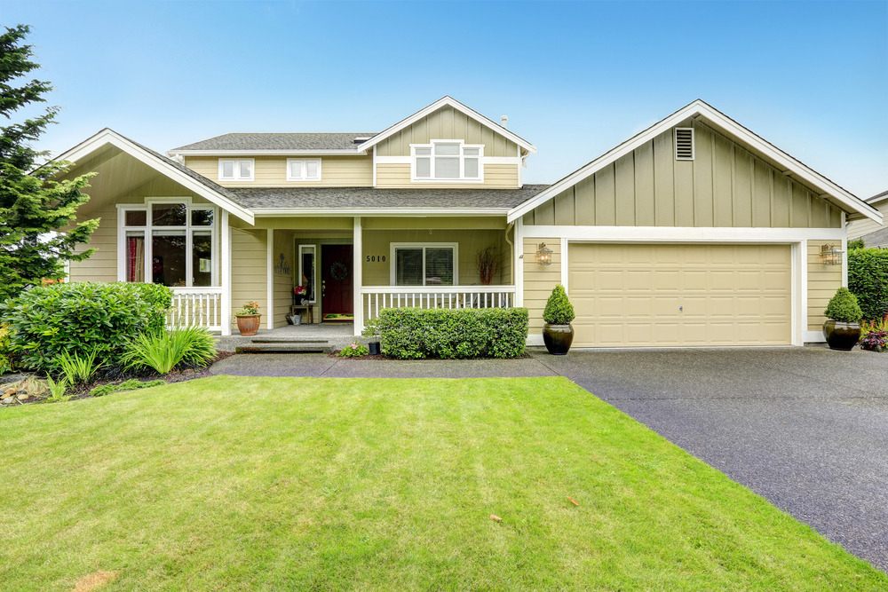 A two-story suburban house with beige siding, a covered front porch, and an attached two-car garage. The yard has green grass, shrubs, and a paved driveway under a clear blue sky.