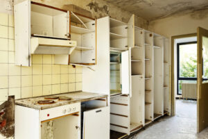A worn-out kitchen with open and damaged white cabinets, a stained electric stove, a partially open refrigerator, yellowed tiles, peeling paint, and a window letting in natural light.