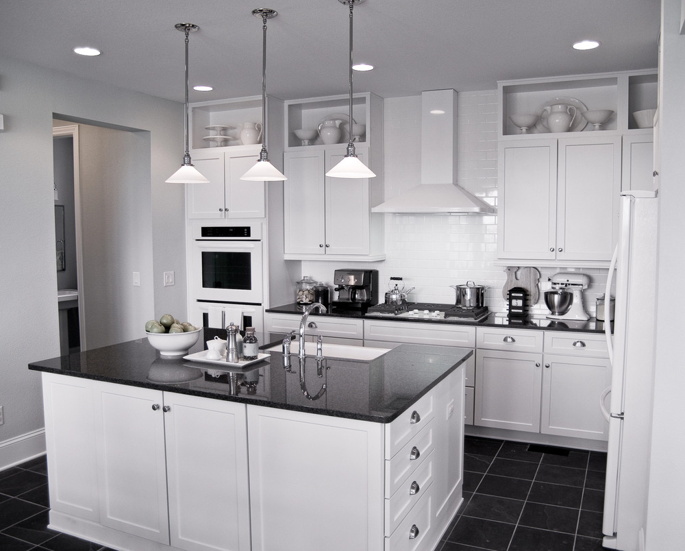 A modern kitchen with white cabinets, a black countertop island, stainless steel appliances, a built-in oven, hanging pendant lights, and open shelves displaying white dishes. The floor is tiled in dark gray.