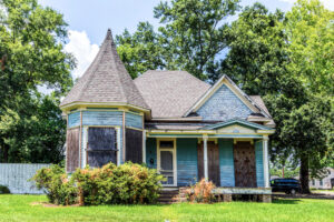 A weathered, blue Victorian-style house with boarded-up windows and a round turret sits on a grassy lawn, surrounded by trees, under a partly cloudy sky. Shrubs grow in front of the porch.
