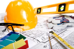 A yellow hard hat, level, safety glasses, work gloves, paint swatches, a tape measure, and a pen rest on top of architectural blueprints.