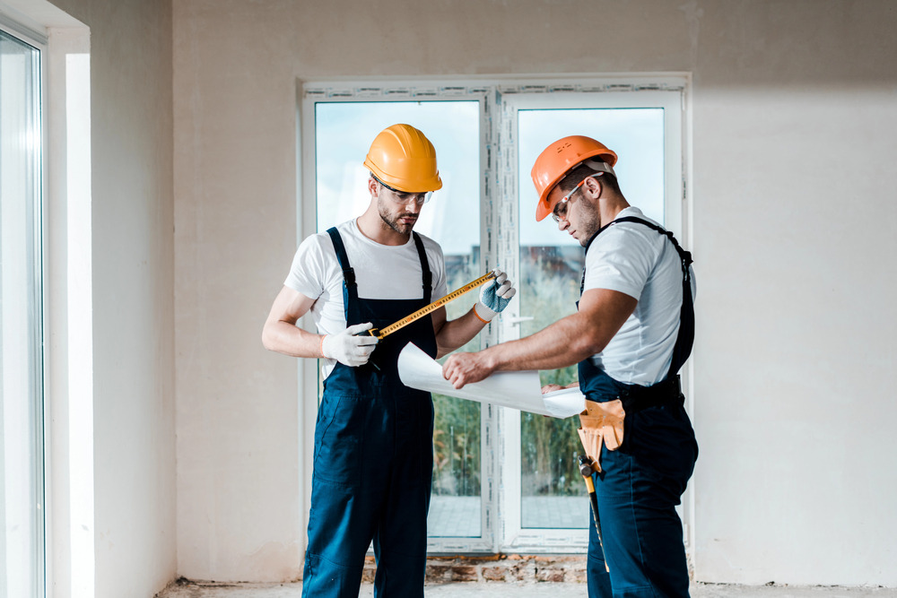 Two construction workers in hard hats and overalls examine a blueprint and measure with a tape measure inside a room with large windows and unfinished walls.