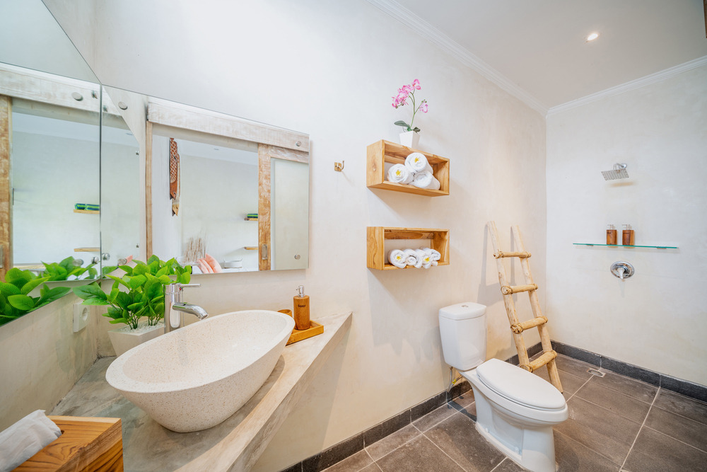 Modern bathroom with a white sink on a stone counter, a large mirror, a toilet, wooden shelves with towels, a potted plant, a decorative ladder, and a glass shelf with toiletries on beige walls and gray tiled floor.