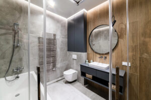 Modern bathroom with gray and wood-textured walls, a wall-mounted toilet, bathtub with shower, a heated towel rail, floating vanity with sink, round mirror, and recessed ceiling lights.