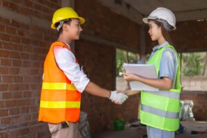 Two construction workers wearing safety vests and helmets shake hands inside an unfinished building, possibly discussing a second story addition cost. One holds documents and a pen, while the other wears gloves. Exposed brick walls and materials are visible.