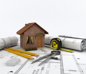 A small model house, a tape measure, rolled architectural blueprints, pencils, an eraser, and a compass lie atop building plans—tools often used to estimate second story addition cost.