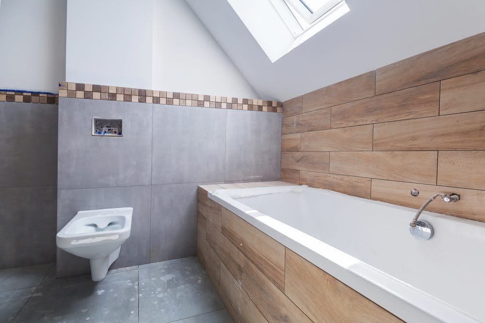 Modern bathroom with a gray tiled floor and wall, a floating white toilet, a rectangular bathtub with wood-look tiles, and a skylight above—ideal inspiration if you’re considering the second story addition cost for extra space.