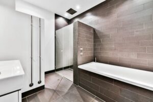Modern bathroom with large rectangular gray tiles, a white bathtub, glass-enclosed shower, wall-mounted towel rail, and a sleek sink. This clean, minimal space adds value to any home and may impact your second story addition cost.