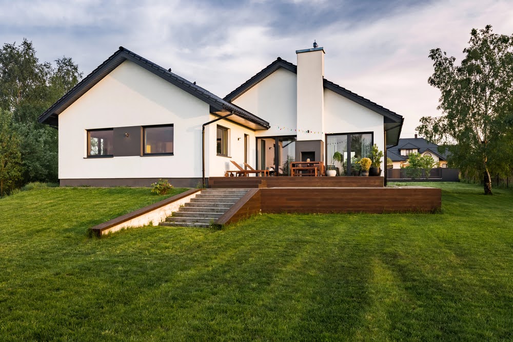 A modern single-story house with white walls and dark roof sits on a well-kept green lawn. The backyard features a raised wooden patio with outdoor furniture—ideal for relaxing as you consider a second story addition cost.