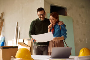 A man and a woman stand together looking at a set of blueprints on a table in a partially finished room, with construction helmets, a laptop, and boxes visible in the background.