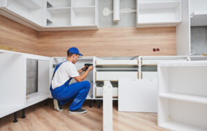 A person wearing blue overalls and a cap is assembling white kitchen cabinets in a room with light wood floors and walls. Tools and cabinet parts are visible around them.