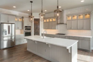 A modern kitchen with light gray cabinets, a large white island with a sink, stainless steel appliances, wooden flooring, pendant lights, and a dark wooden door in the background.