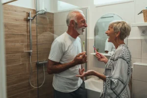 Two older adults stand in a modern bathroom holding toothbrushes, smiling and talking to each other. A shower with a glass door and a mirror with a sink are visible in the background.