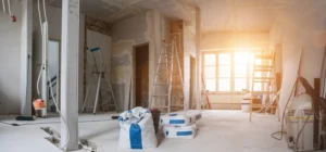 A room under renovation with exposed walls, ladders, construction materials, and tools scattered around; sunlight streams through a large window in the background.